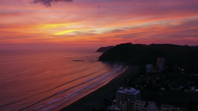 Aerial: Gentle Waves Crashing on Beach During Colorful Tropical Sunset and Silhouette of Jaco and Mountains