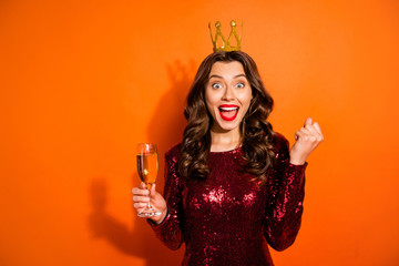 Portrait of excited bachelor girl having crown on her head hold champagne hear news about her win on prom party scream wow omg wear burgundy red dress isolated over orange color background