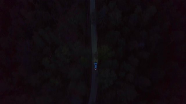 Aerial view white premium car driving on dark countryside road among summer forest at night. View from drone above car with xenon headlights riding on night road through green forest