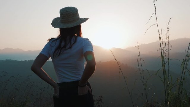 A young woman in a hat with hands in the back pocket watching the beautiful sunrise or sunset over high mountains in the distance. Soft warm lights as tall grass moving in the wind.   
