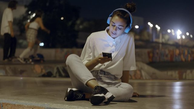 Beautiful Female Listening To Music on Wireless Headphones dancing During Training In city skate Park