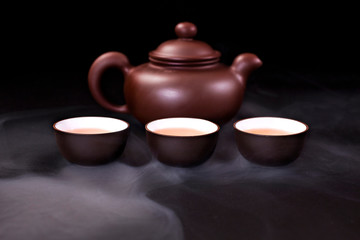 Chinese clay teapot and ceramic tea cups with vapor on a black background.