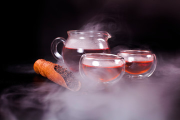 Glass tea cups, glass jug of tea, black tea in a scoop with vapour on a black background.