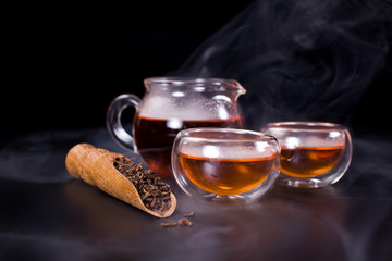 Glass tea cups, glass jug of tea, black tea in a scoop with vapour on a black background.