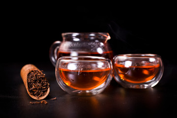 Glass tea cups, glass jug of tea with black tea in a scoop on a black background.