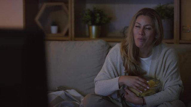 Dolly-out Shot Of Happy Woman Sitting Cross-legged On Couch In Dark Living Room And Smiling While Eating Chips And Watching TV