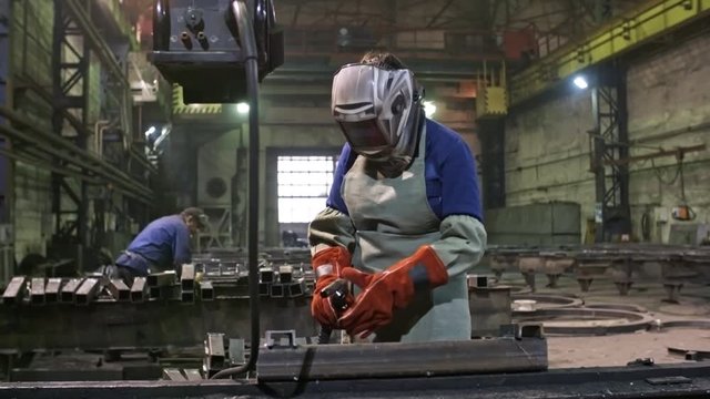 Tracking shot shot of female worker in gloves putting on safety helmet and welding steel part at metal fabrication facility