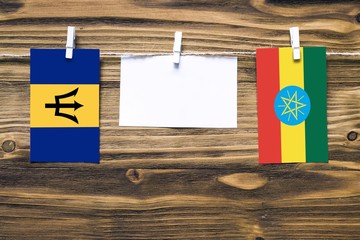 Hanging flags of Barbados and Ethiopia attached to rope with clothes pins with copy space on white note paper on wooden background.Diplomatic relations between countries.