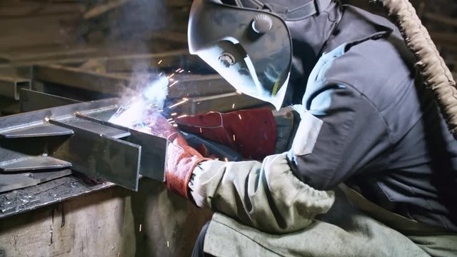 Handheld shot of unrecognizable male worker in protective uniform and helmet welding steel beams at metal fabrication facility