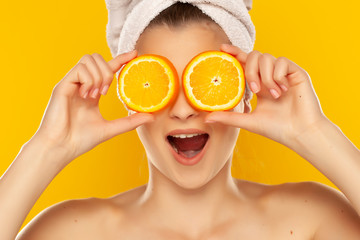 Young beautiful woman with towel on her head holding slices of oranges in front of her eyes on yellow background