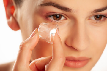 Portrait of young beautiful holding ice cube under her eye on white background
