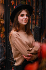 Autumn girl stands near a fence with red ivy. Sweater, hat and leather skirt. Atmosphere.