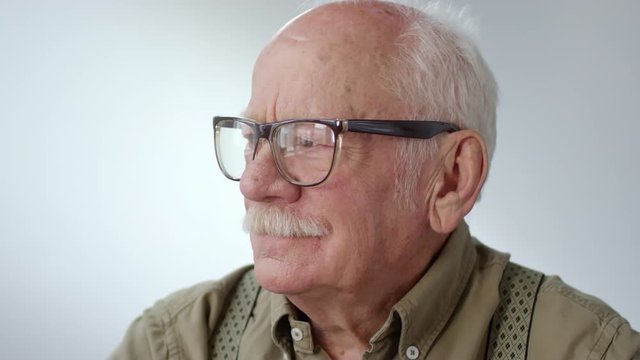Portrait shot with close up of charismatic senior man with grey hair and mustache posing isolated on grey background. He is gazing into distance, then taking off his glasses and looking at camera