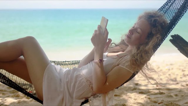 Relaxed woman browsing smartphone while lounging in hammock on beach