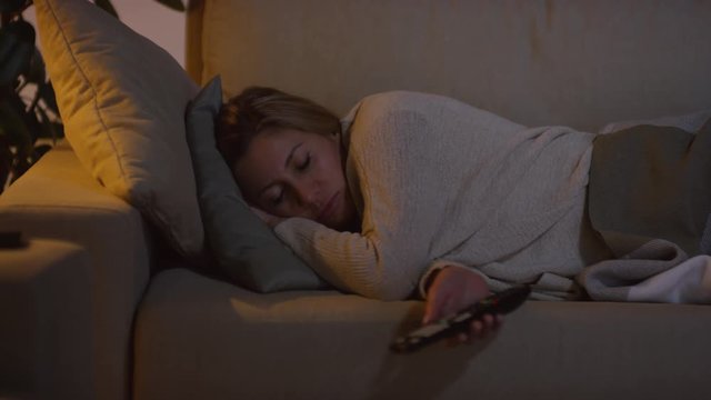 Tracking of woman holding remote control and sleeping on couch in dark living room with TV on