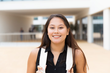 Happy excited female tourist posing outside. Young mix raced woman in casual standing near city building, holding backpack straps, looking at camera, smiling. Young woman portrait concept