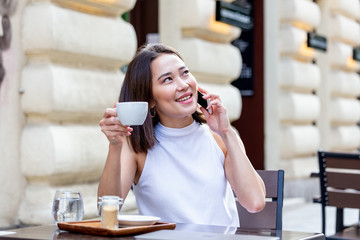 Young Asian woman sitting in coffee shop at wooden table, drinking coffee and using smartphone.On table is laptop. Girl browsing internet, chatting, blogging.