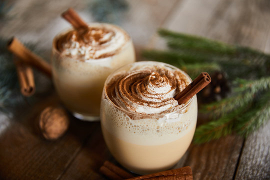 selective focus of eggnog cocktail with whipped cream near cinnamon sticks, spruce branch and walnuts on wooden table