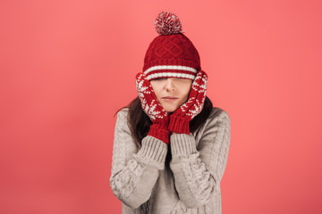 woman in red gloves touching face and covering eyes with knitted hat on pink