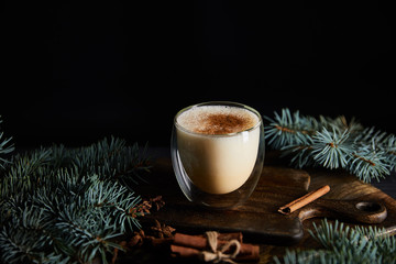 delicious eggnog cocktail on cutting board near spruce branches and cinnamon sticks isolated on black