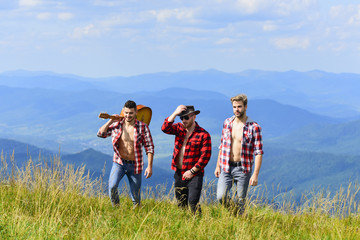 Friendly guys with guitar hiking on sunny day. Tourists hiking concept. Enjoying freedom together. Group of young people in checkered shirts walking together on top of mountain. Hiking with friends
