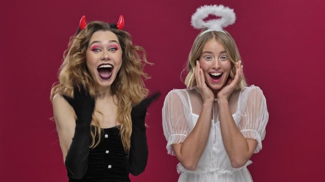 Angel and demon girls pleasantly surprised happy and smile in carnival costumes isolated over red wall background