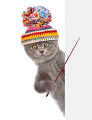 Cat wearing a warm hat peeks and points on empty space banner. isolated on white background