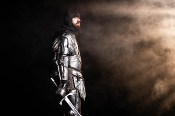 handsome knight in armor holding sword on black background - obrazy, fototapety, plakaty