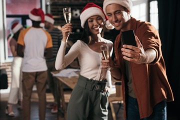 selective focus of cheerful businessman and businesswoman in santa hats taking selfie on smartphone while holding champagne glasses