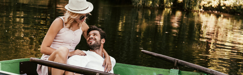 panoramic shot of young woman in sundress and hat hugging happy boyfriend while sitting in boat