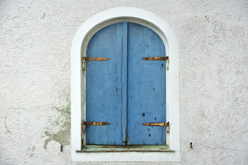 An old weathered blue shutter closes an old window with round arches in an old wall