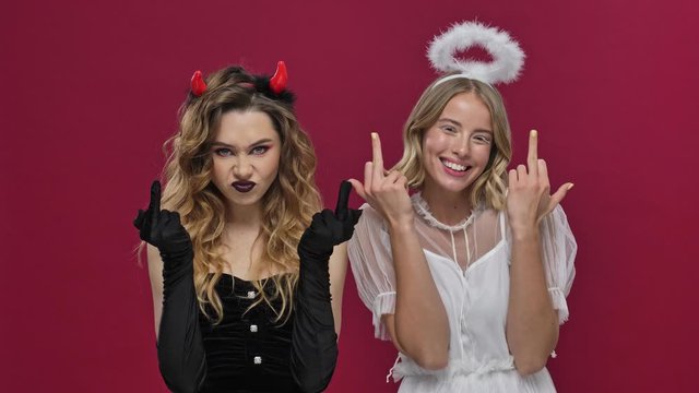Happy angel and angry demon girls showing middle finger and smiling in carnival costumes isolated over red wall background