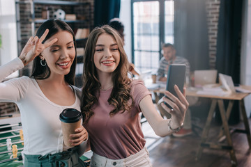 young, cheerful businesswoman taking selfie on smartphone with cheerful colleague holding coffee to go and showing victory gesture