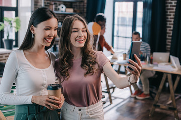 young, cheerful businesswoman taking selfie on smartphone with cheerful colleague holding coffee to go