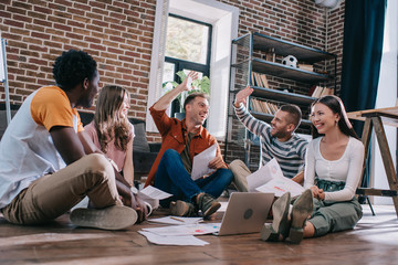 happy businessmen giving high five while sitting on floor near young multicultural colleagues