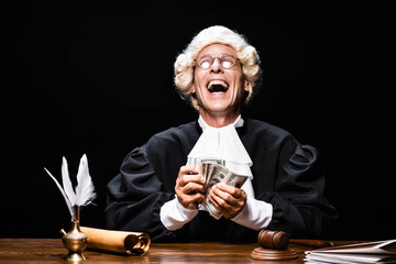 smiling judge in judicial robe and wig sitting at table and holding money isolated on black