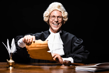 smiling judge in judicial robe and wig sitting at table and holding document isolated on black