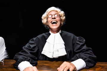 smiling judge in judicial robe and wig sitting at table isolated on black
