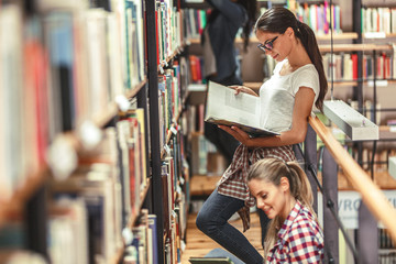Two female students reads a book by the bookshelf at the library. Reading a book.	