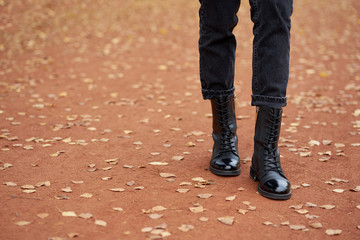 Woman's legs in fashionable autumn leather black boots and trendy black jeans. Trendy youth casual outfit for everyday. Modern street collection. Closeup. Autumn road with yellow leaves on background.