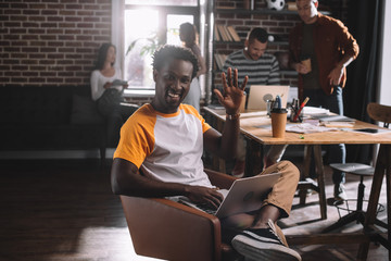 smiling african american businessman using laptop while sitting in armchair and waving hand near...