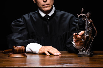 cropped view of judge in judicial robe sitting at table with gavel and themis figure isolated on black