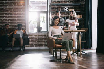 attractive businesswoman sitting in armchair and using laptop near young multicultural colleagues