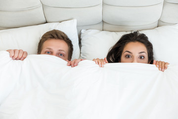 young couple handsome husband and his beautiful wife cuddling in bed in bright bedroom with blanket covering half of their faces family morning idyll