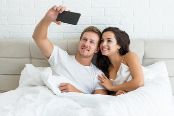 young happy couple handsome husband and his beautiful wife just woke up in bed shooting selfie on smartphone early in the morning social media addiction