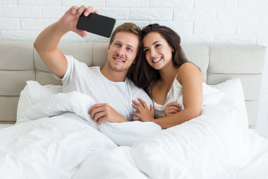 young couple handsome husband and his beautiful wife just woke up in bed shooting selfie on smartphone early in the morning social media addiction