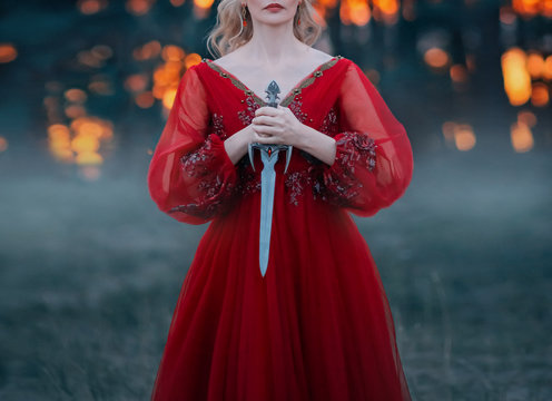 A mysterious blond woman holds a dagger of beautiful Gothic design in her hands. Fantasy lady soldier fashion warrior in a dark forest. Shooting without a face, only lips and hands. Cut off the head