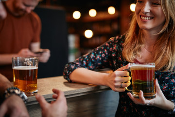 Smiling beautiful blonde leaning on bar counter, holding pint of beer and talking to her friend. Nightlife.