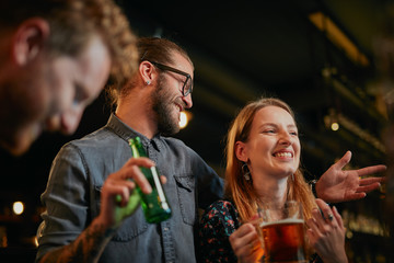 Handsome caucasian man with eyeglasses hugging his girlfriend and talking to her while standing in a pub and drinking beer. Nightlife.