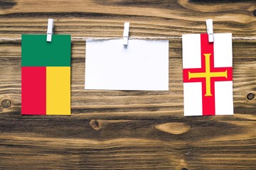 Hanging flags of Benin and Guernsey attached to rope with clothes pins with copy space on white note paper on wooden background.Diplomatic relations between countries.